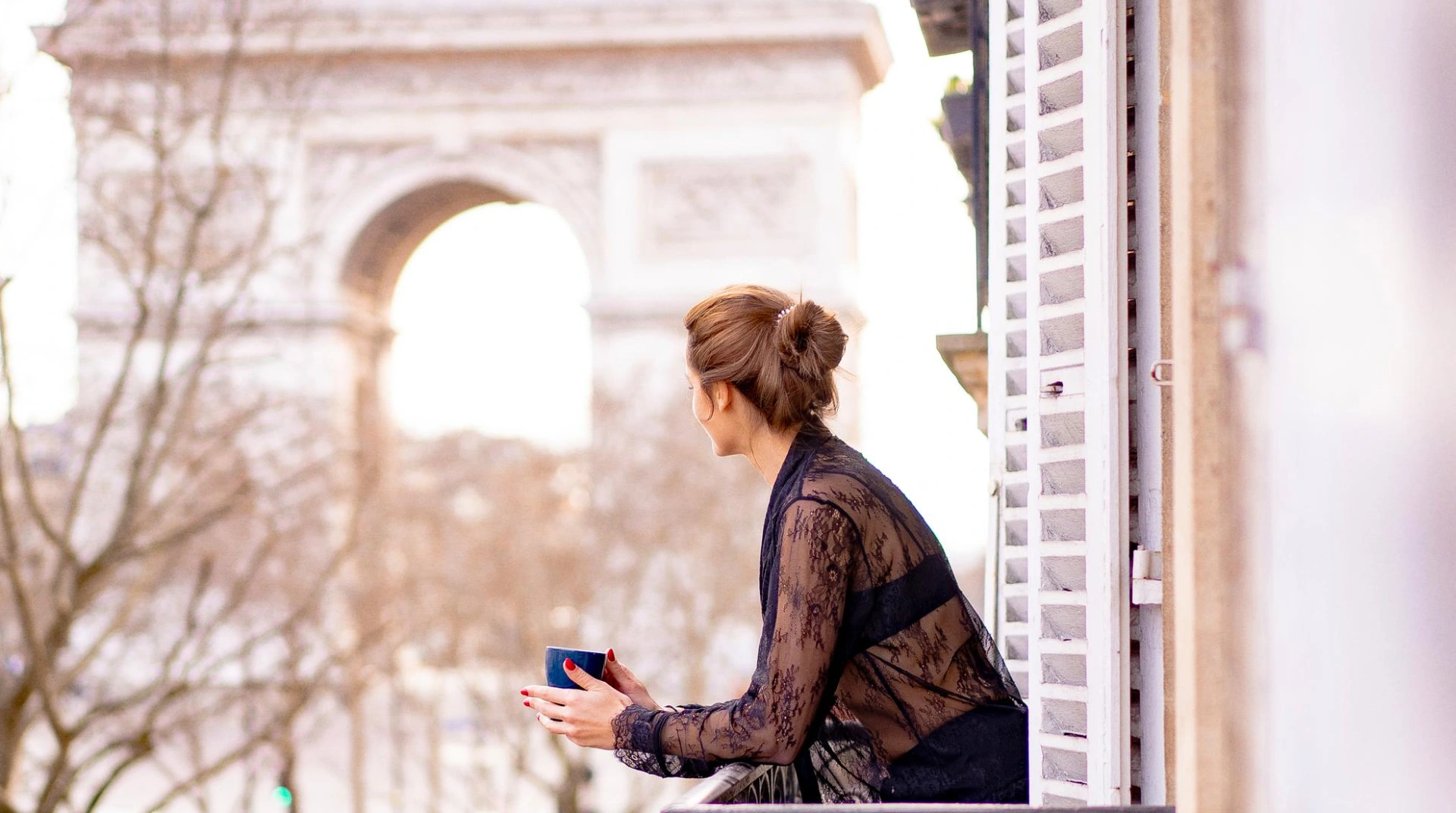Femme de dos au balcon, tenant une tasse, avec vue sur l'Arc de Triomphe.