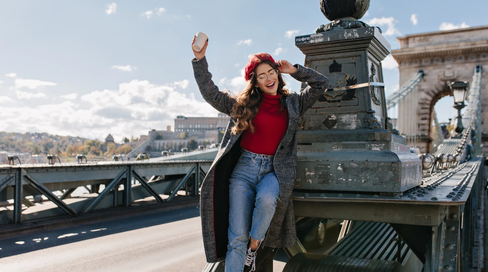 Femme en manteau sur un pont, souriante, tenant un gobelet et portant un béret rouge.