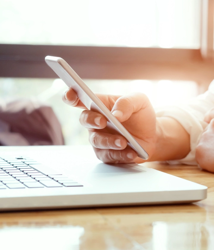Personne tenant un smartphone au-dessus d'un ordinateur portable sur une table en bois.