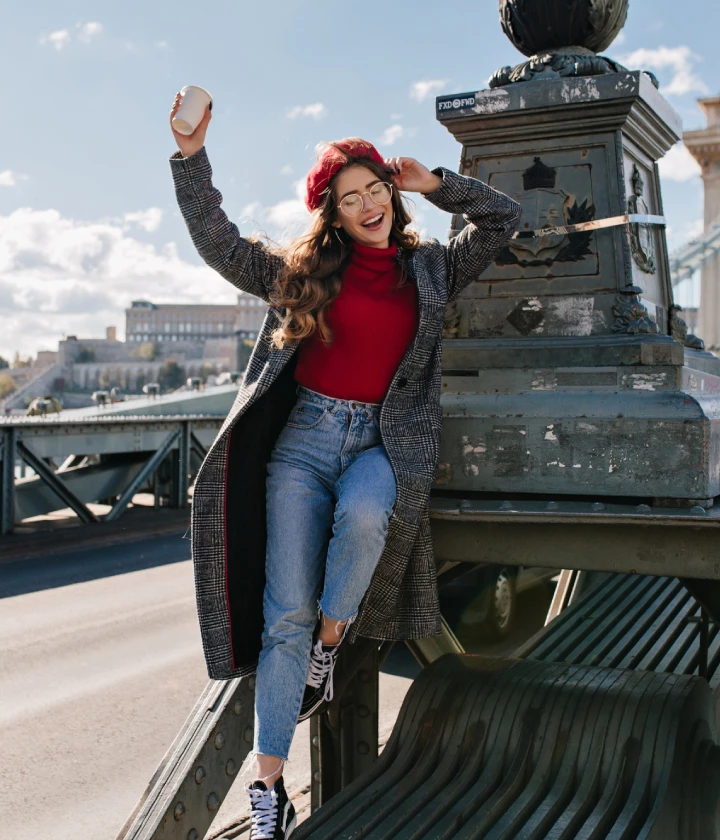 Femme souriante en manteau et béret rouge, lève une tasse sur un pont ensoleillé.