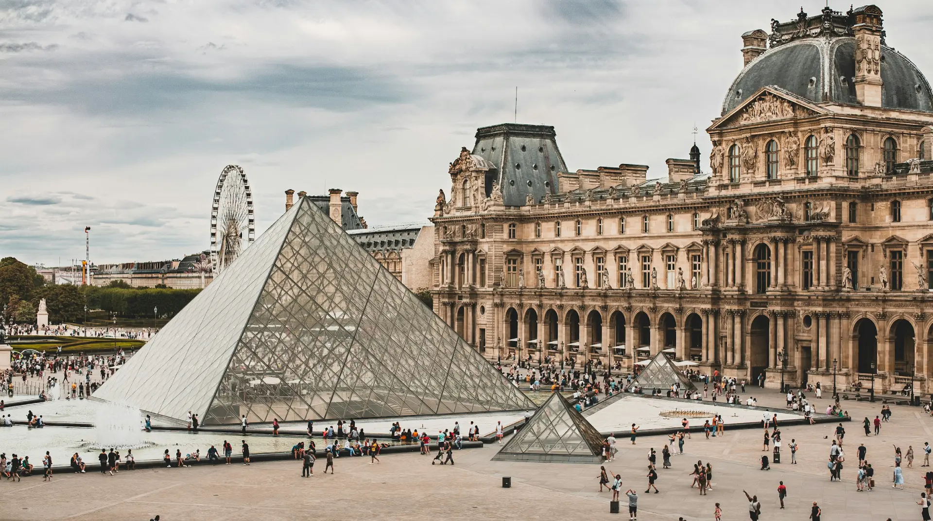Pyramide de verre du Louvre avec grande roue en arrière-plan, nombreux visiteurs.