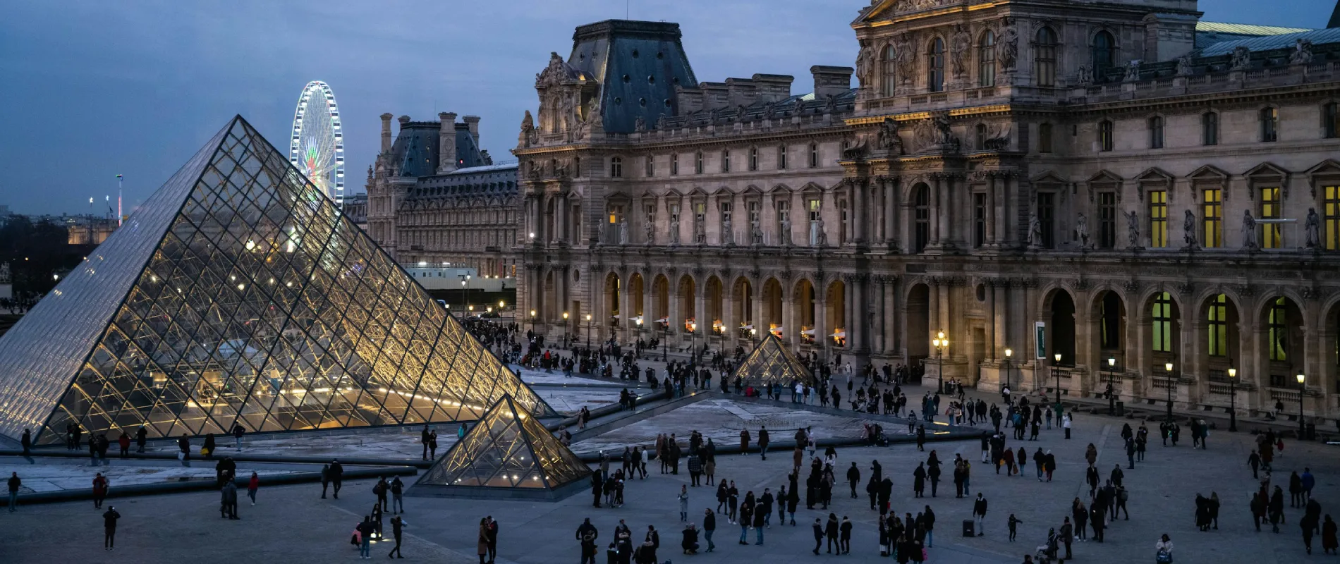 Cour du Louvre au crépuscule, pyramide illuminée, grande roue en arrière-plan.