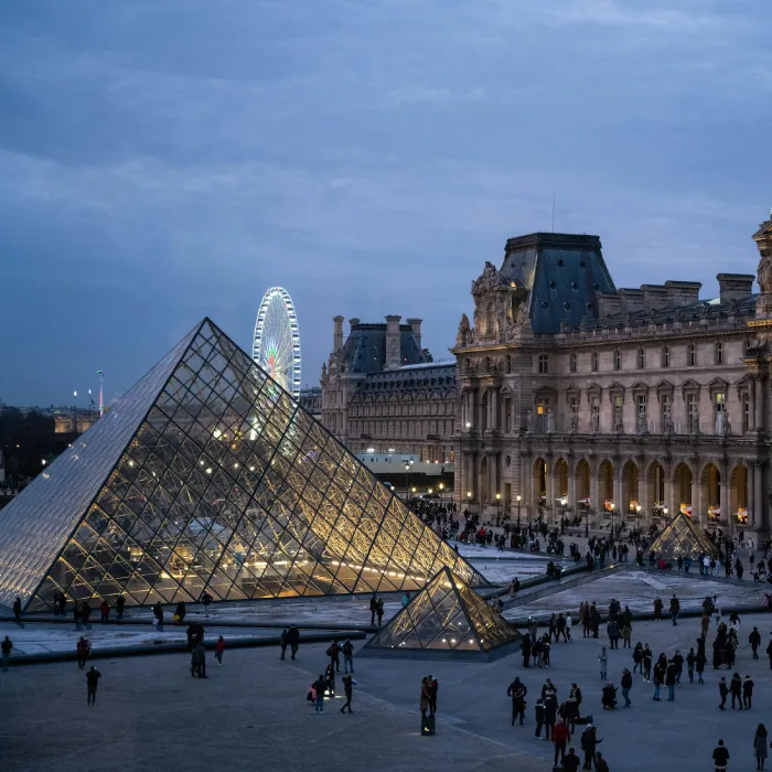 La pyramide du Louvre illuminée, avec la grande roue en arrière-plan, sous un ciel du soir.
