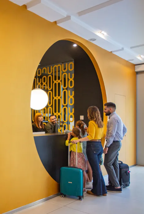 Familia en recepción de hotel moderno, con equipaje, frente a mostrador amarillo con diseño geométrico.