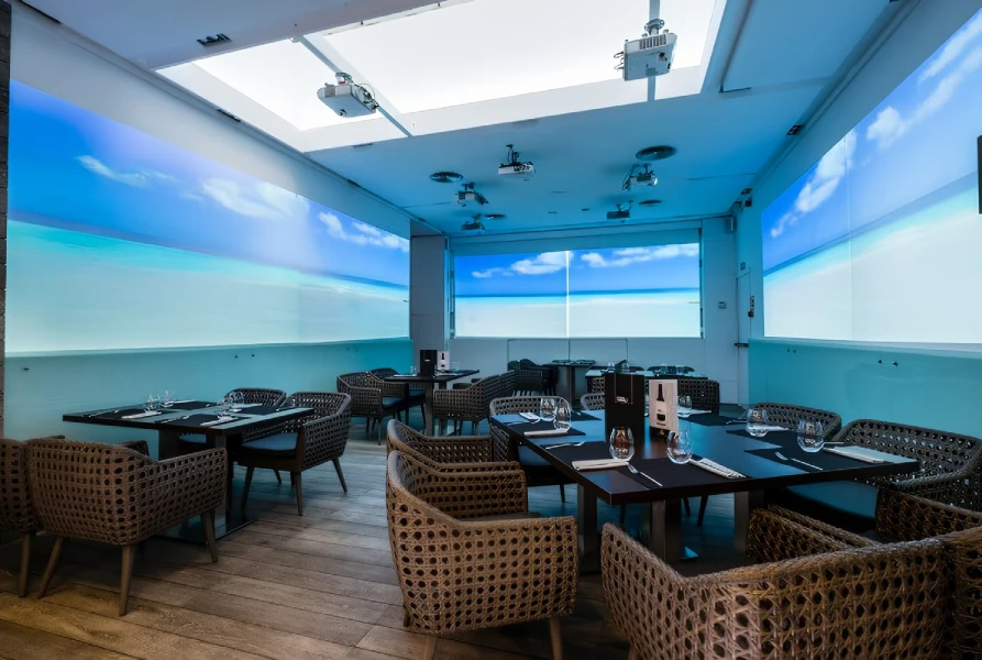 Restaurante moderno con sillas tejidas y pantallas mostrando una playa en las paredes.
