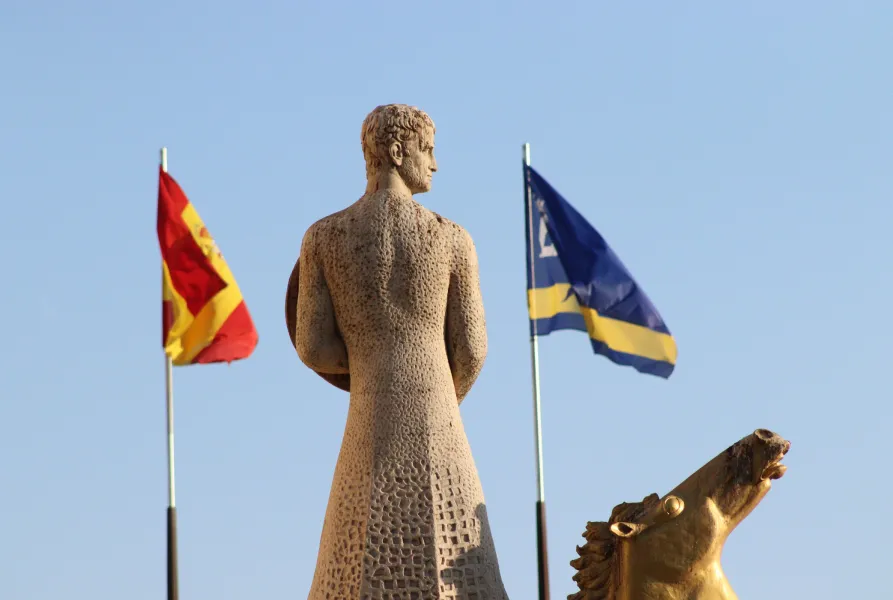Una estatua de espaldas junto a banderas roja-amarilla y azul-amarilla sobre un cielo despejado.