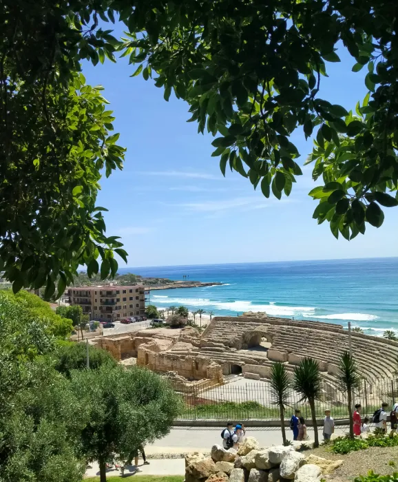 Vista del mar tras un antiguo anfiteatro romano rodeado de vegetación y edificios modernos.