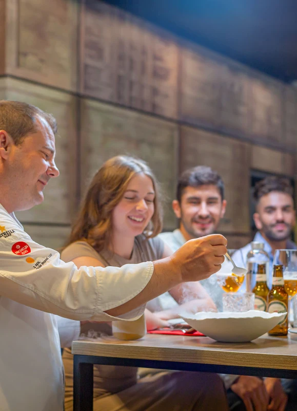 Chef sirviendo comida a tres personas sentadas en una mesa con cervezas.