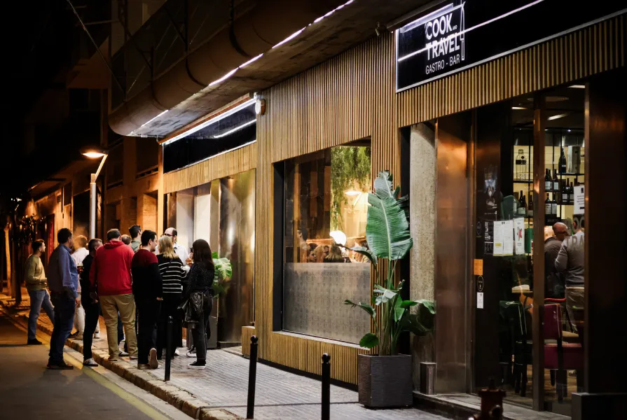 Personas esperan afuera de un restaurante elegante con un letrero que dice "Cook... Travel".