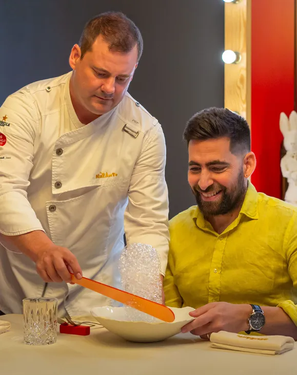 Chef preparando un platillo burbujeante frente a un hombre sonriente en un restaurante.
