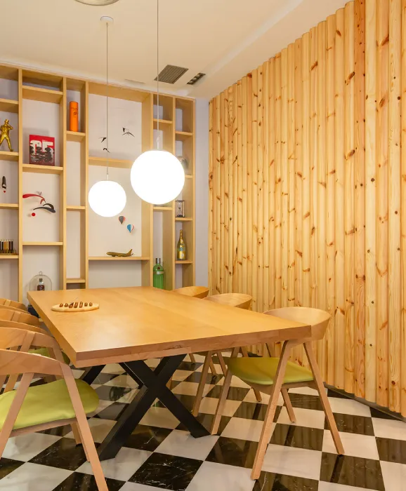 Sala de comedor con mesa de madera, sillas, lámparas colgantes y estanterías decorativas al fondo.