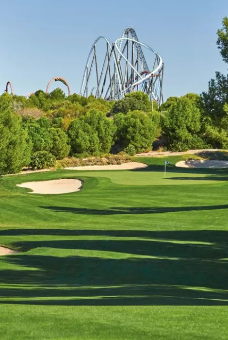 Campo de golf con colinas verdes y un banderín, con montañas rusas visibles al fondo entre árboles.
