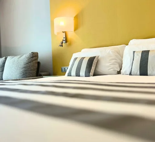 Cama con almohadas y cojines, pared amarilla, lámpara encendida. Ambiente acogedor.