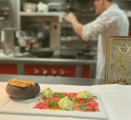 Plato gourmet decorado y chef cocinando en el fondo de una cocina moderna.