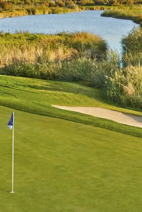 Campo de golf con bandera, césped, búnker y lago al fondo.
