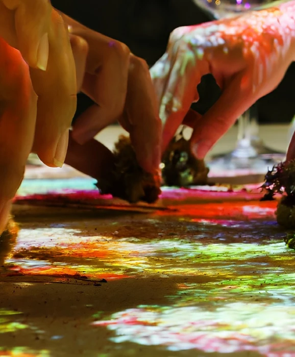 Manos sobre una mesa iluminada con luces de colores, cerca de una copa de vino.