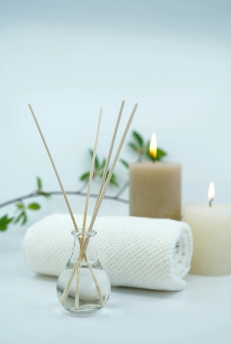 Difusor aromático, velas encendidas y toalla blanca sobre fondo blanco con plantas.
