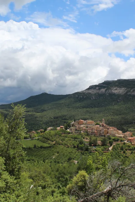 Pueblo en una colina rodeado de montañas y vegetación bajo un cielo parcialmente nublado.