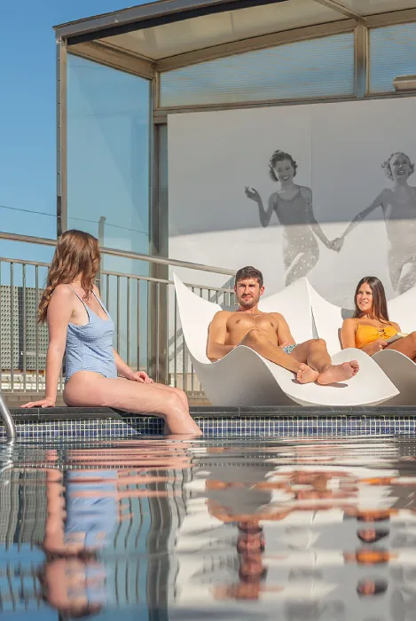 Personas relajándose junto a una piscina, con sillas modernas y un mural al fondo.