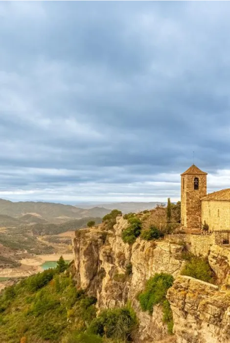 Iglesia en un acantilado rocoso con un paisaje montañoso bajo un cielo nublado.
