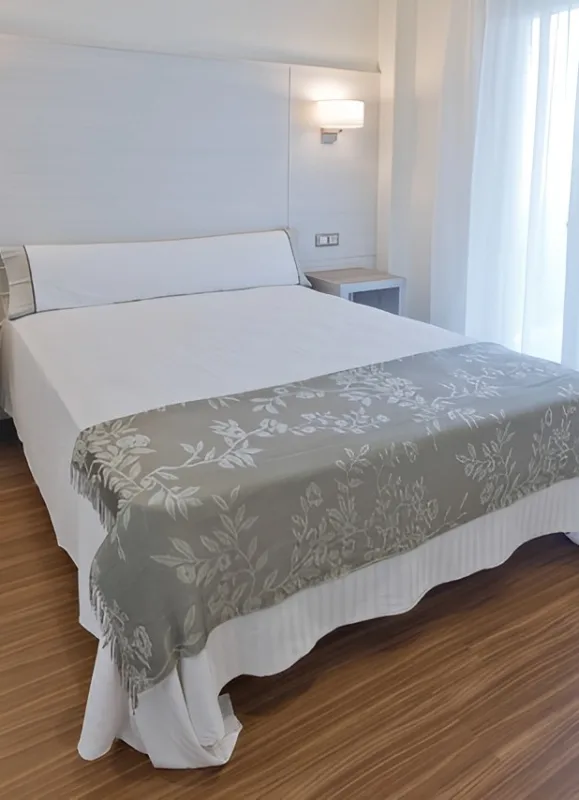 Cama blanca en habitación luminosa, con manta gris decorativa, suelo de parquet y luz suave.