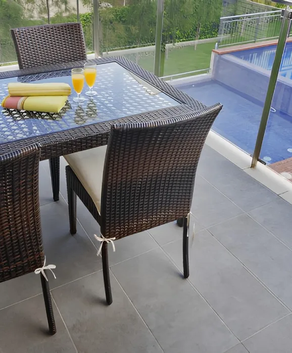Juego de mesa y sillas de mimbre en terraza, dos copas de jugo y toallas cerca de una piscina.