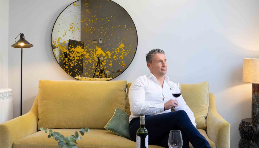 Hombre en sofá mostaza, espejo redondo con manchas amarillas, copa de vino.