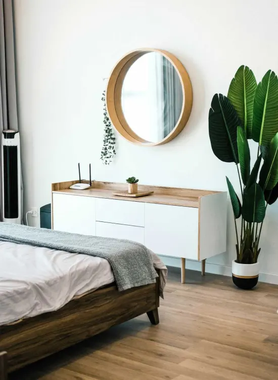 Dormitorio minimalista con espejo redondo, cómoda y planta decorativa junto a la cama.