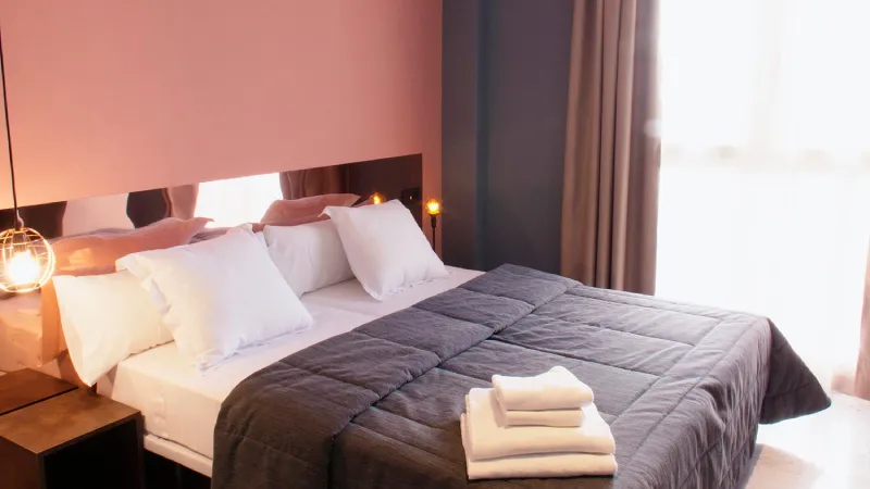 Cama en habitación luminosa con almohadas y colcha oscura.