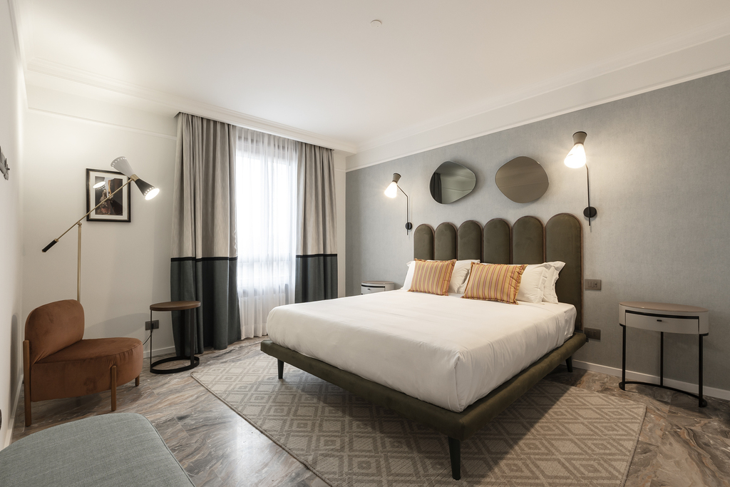Habitación moderna con cama doble, sillón, lámparas y ventanas con cortinas.