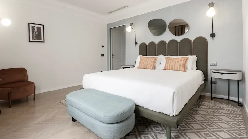 Habitación moderna con cama grande, dos mesitas, espejo y bancos acolchados al pie de la cama.