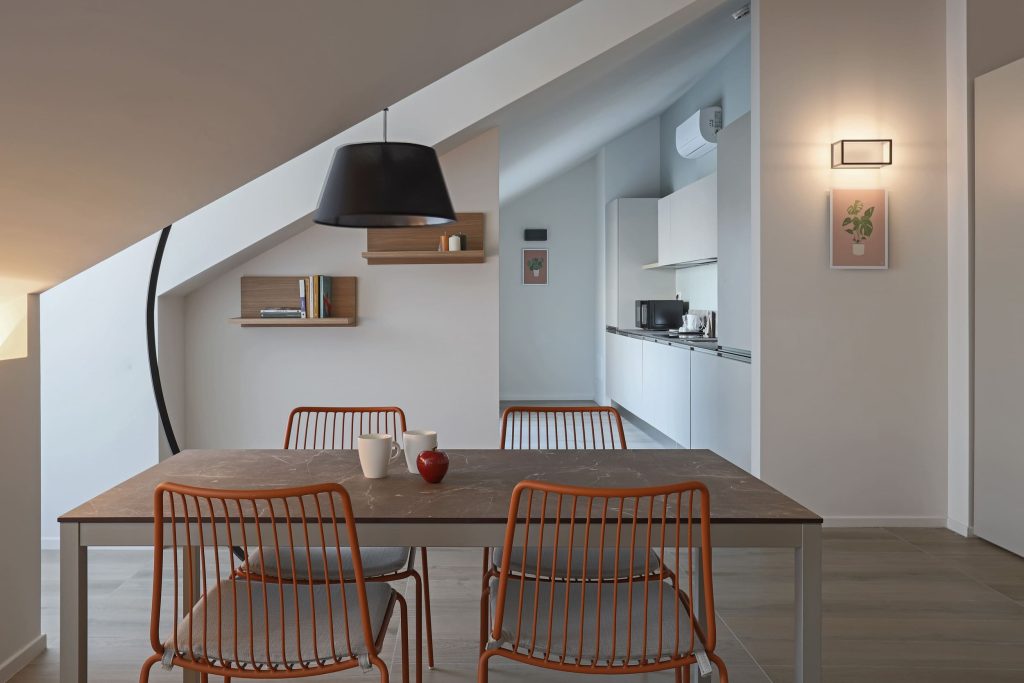 Comedor moderno con sillas naranjas y cocina blanca al fondo.