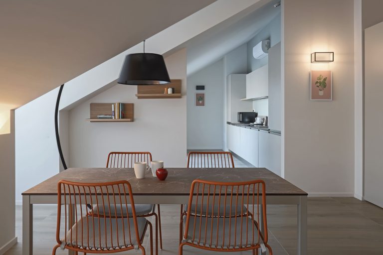 Comedor moderno con sillas naranjas y cocina blanca al fondo.