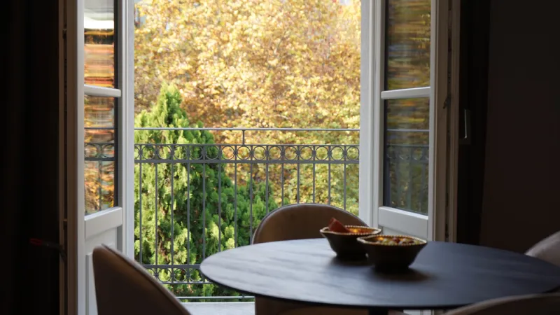 Vista de una terraza con árboles otoñales desde una habitación con mesa redonda.