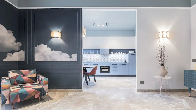 Sala moderna con sillón geométrico, cocina al fondo y paredes decoradas con nubes y lámparas.
