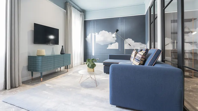 Sala moderna con sofá azul, televisor, mesa de centro y mural de nubes en la pared.