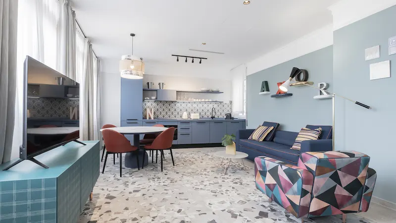 Sala de estar moderna con cocina azul, sofá, silla geométrica y comedor. Suelo de terrazo.