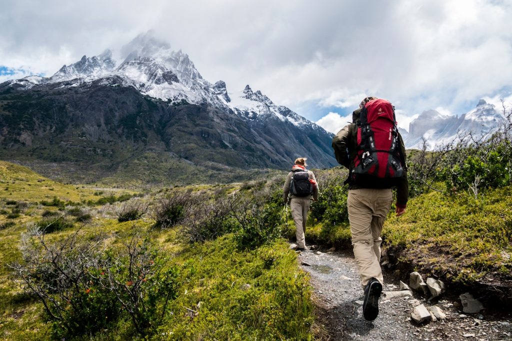 Dos personas hacen senderismo en un paisaje montañoso con montañas nevadas y cielo parcialmente nublado.