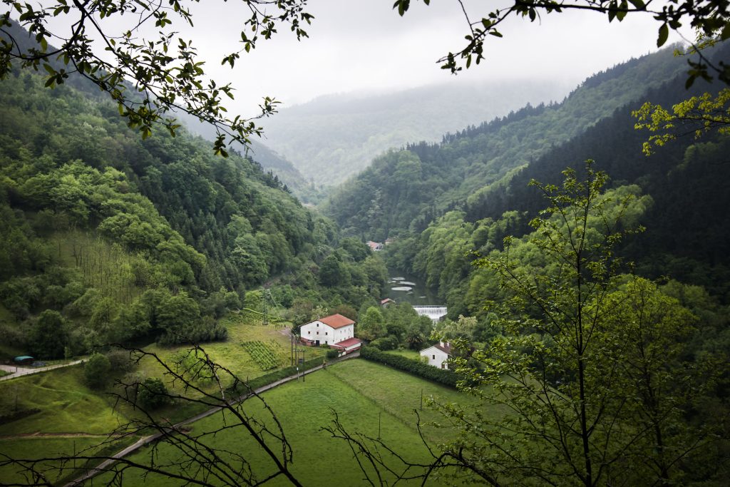 Valle verde con casas blancas, rodeado de colinas boscosas y bajo un cielo nublado.