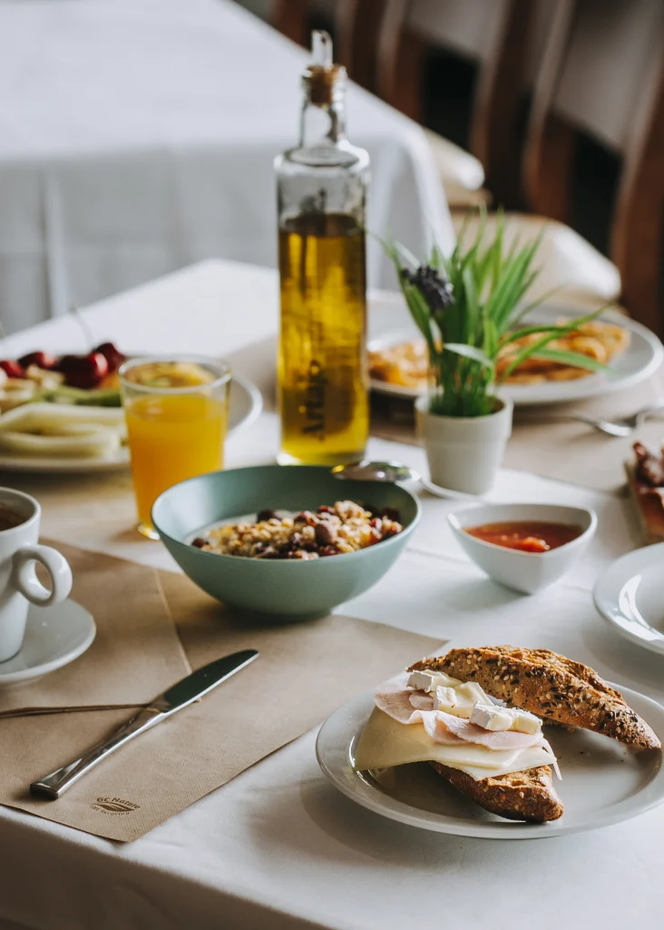 Mesa con desayuno: jugo, croissant, tazón de cereal, café, botella de aceite y planta pequeña sobre mantel blanco.