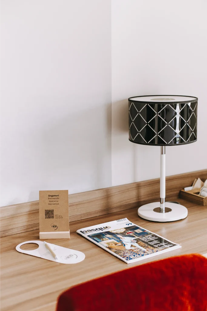 Lámpara con pantalla negra sobre mesa de madera, revista, cartel de bienvenida y colgador de puerta.