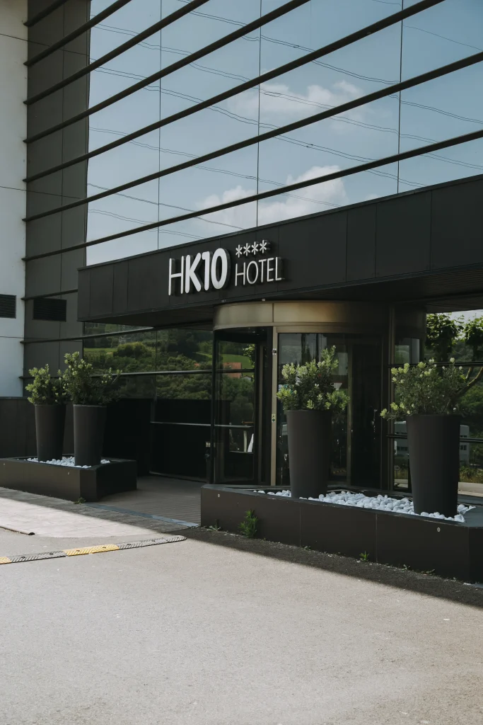 Entrada del hotel HK10 con fachada de vidrio, plantas en macetas negras y un letrero cuatro estrellas.