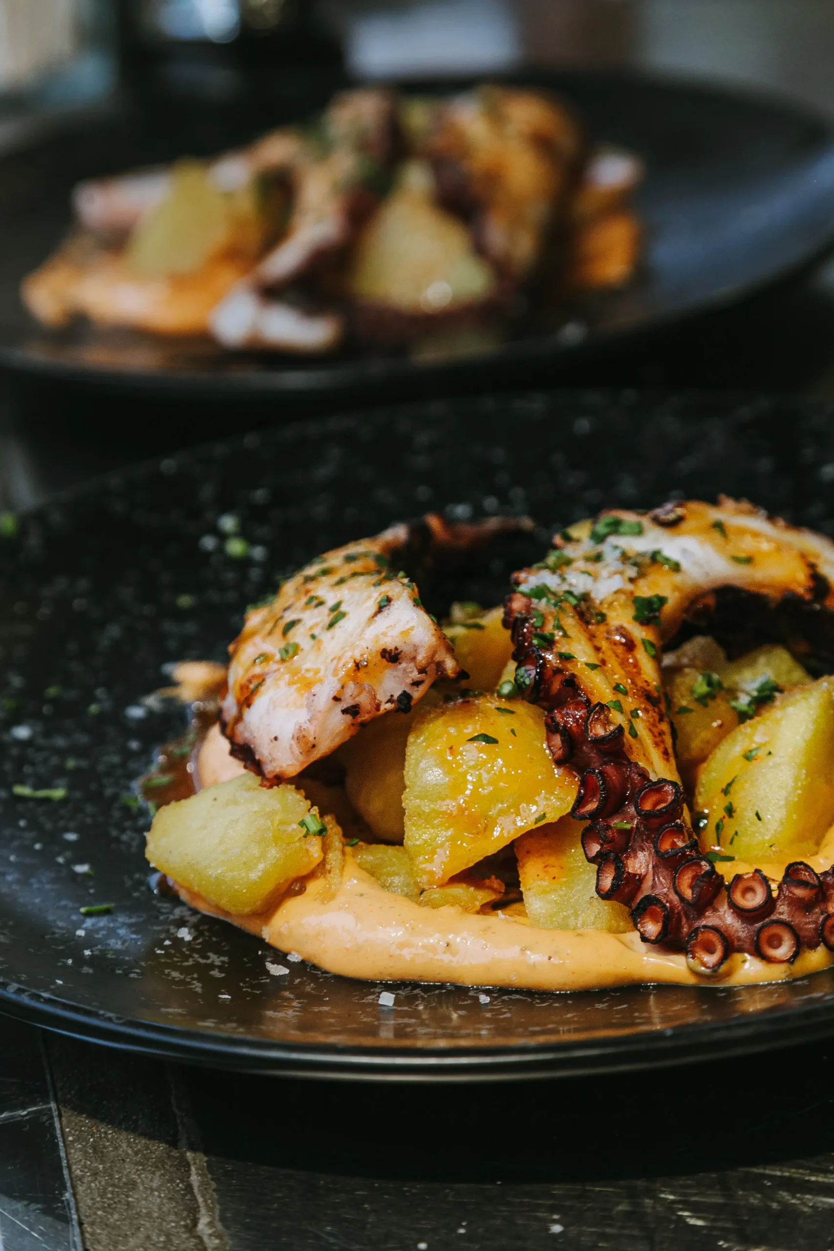 Plato negro con pulpo asado, papas doradas y salsa, espolvoreado con hierbas.