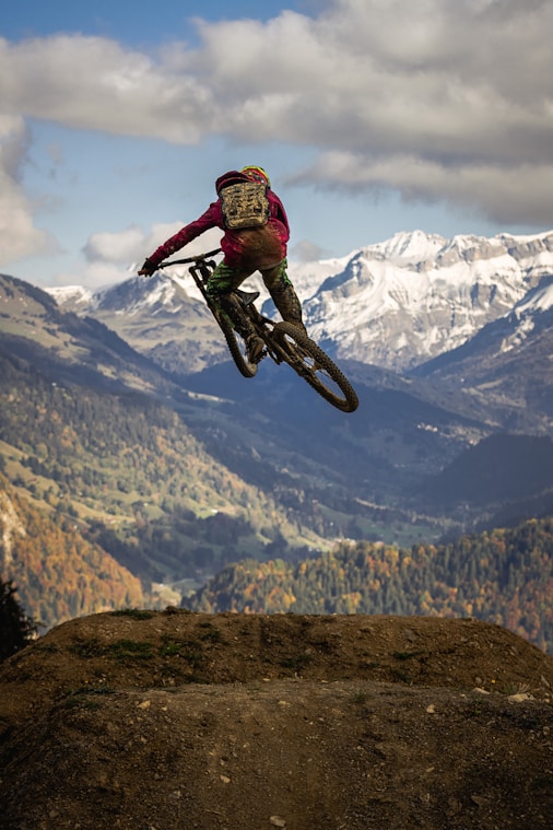 Persona haciendo un salto en bicicleta de montaña en un paisaje alpino con montañas nevadas al fondo.