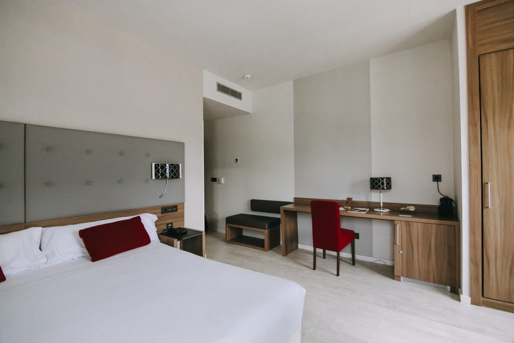 Habitación de hotel moderna con cama, escritorio de madera, silla roja y lámpara de mesa, ambiente sencillo y acogedor.