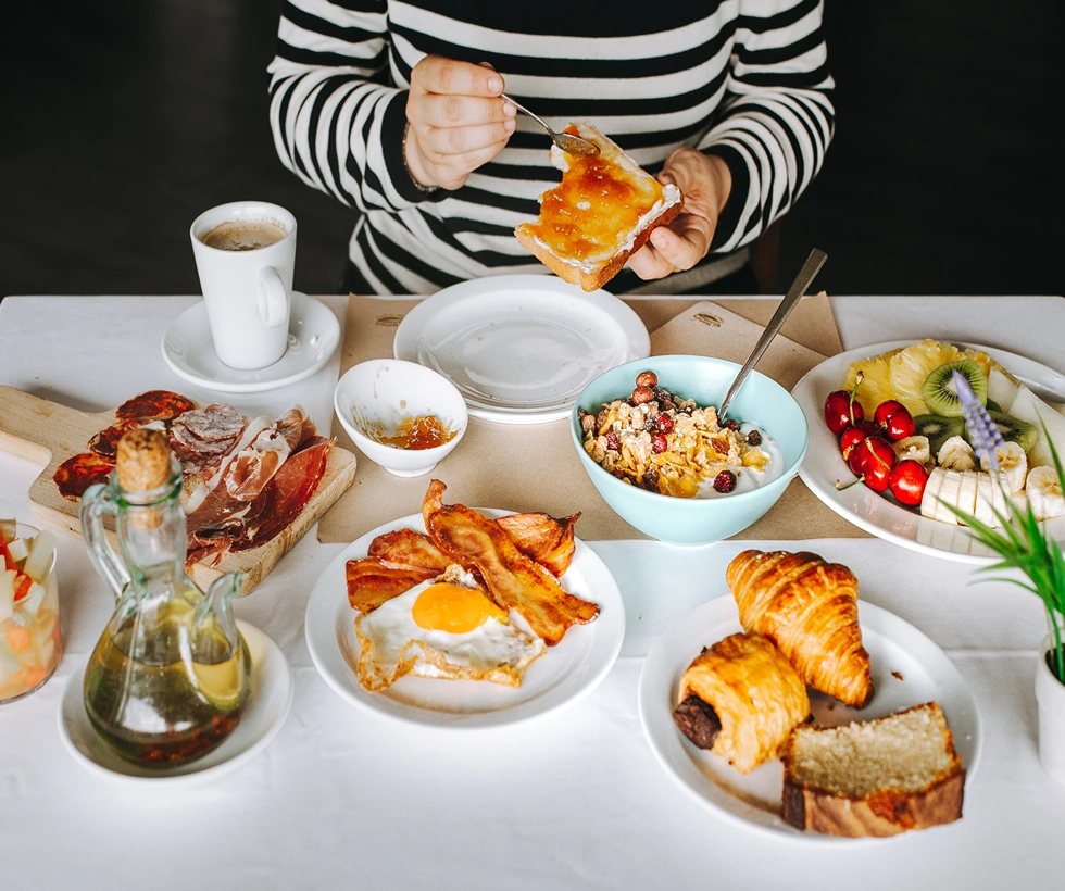 Desayuno variado: tostada con mermelada, croissants, huevo con tocino, frutas, cereales, café y embutidos.
