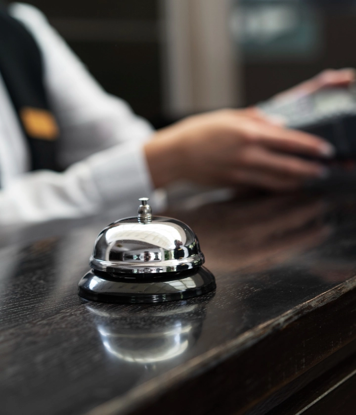 Campana de hotel en un mostrador de recepción, persona de uniforme usando dispositivo electrónico al fondo.