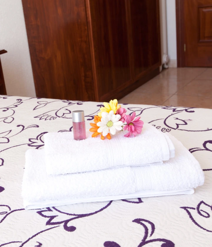 Toallas blancas con flores y frasco sobre una cama con colcha de diseño floral.