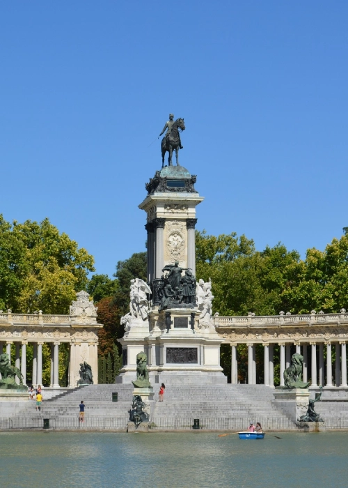 Monumento ecuestre rodeado de columnas junto a un lago, con personas y bote en el agua.