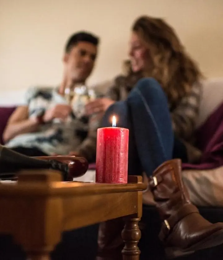 Pareja sentada en un sofá, desenfocada, con una vela roja encendida en primer plano sobre la mesa.